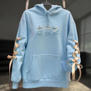 May include: Light blue hoodie with a drawstring hood and a kangaroo pocket. The sleeves feature gold ribbon lacing. The front of the hoodie has an embroidered design of three cartoon characters. The hoodie is hanging on a black hanger.
