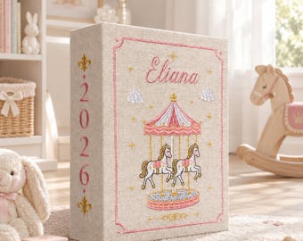 May include: A rectangular beige fabric-covered memory book featuring embroidered pink and gold details. The front displays the name Eliana, a carousel with two horses, and small clouds. The spine is embroidered with the year 2026 in pink.