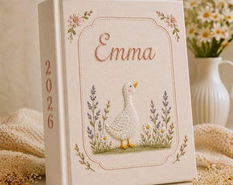 May include: A cream-colored fabric-covered book featuring embroidered details. The front cover displays the name Emma, a white duck, and floral motifs. The spine is embroidered with the year 2026. The design includes pink, purple, and green thread.