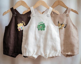 May include: Three baby rompers in brown, white, and tan, hanging on a wooden rod. Each romper features a different embroidered cartoon character and name: brown with "Travis", white with "Lucas", and tan with "Violet". The rompers have button closures and a simple design.