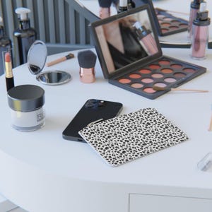 May include: A black smartphone with a white and black animal print phone case on a white table with makeup brushes, a compact mirror, and a makeup palette.