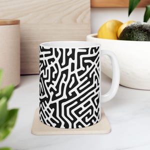 May include: White ceramic mug featuring a black and white abstract design. The pattern resembles a maze or labyrinth.
