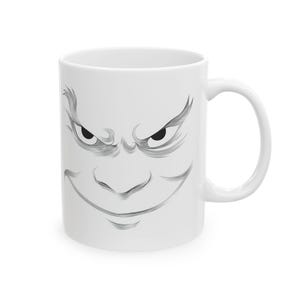 Peut inclure: Mug en céramique blanche avec une illustration en noir et gris d'un visage en colère. Le design comprend des sourcils froncés, des yeux plissés et un large sourire sinistre. La tasse a une anse incurvée.