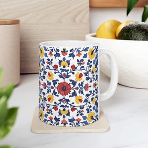 May include: White ceramic mug with a blue, yellow, and red floral pattern. The mug has a handle and is sitting on a beige coaster.