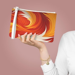 May include: A rectangular clutch with a white wrist strap and zipper. The clutch features a vibrant abstract design in shades of red, orange, and yellow, with white accents. The clutch is held up by a hand.