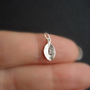 May include: A silver leaf charm with the letter L engraved on it. The charm is hanging from a silver jump ring.