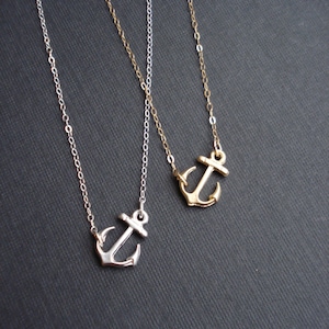 May include: Two delicate anchor necklaces, one silver and one gold, on a dark gray background. The anchors are small and detailed.