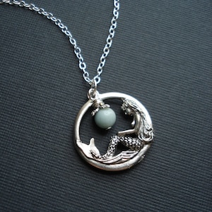 May include: Silver necklace with a mermaid pendant and a light blue gemstone. The mermaid is sitting on a crescent moon.