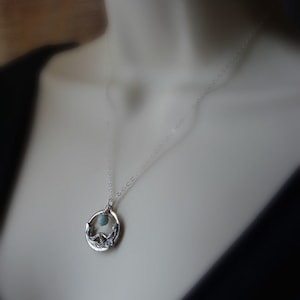 May include: A silver necklace with a delicate chain and a pendant featuring a circular design with a small blue gemstone in the center. The pendant is detailed with a stylized floral motif.