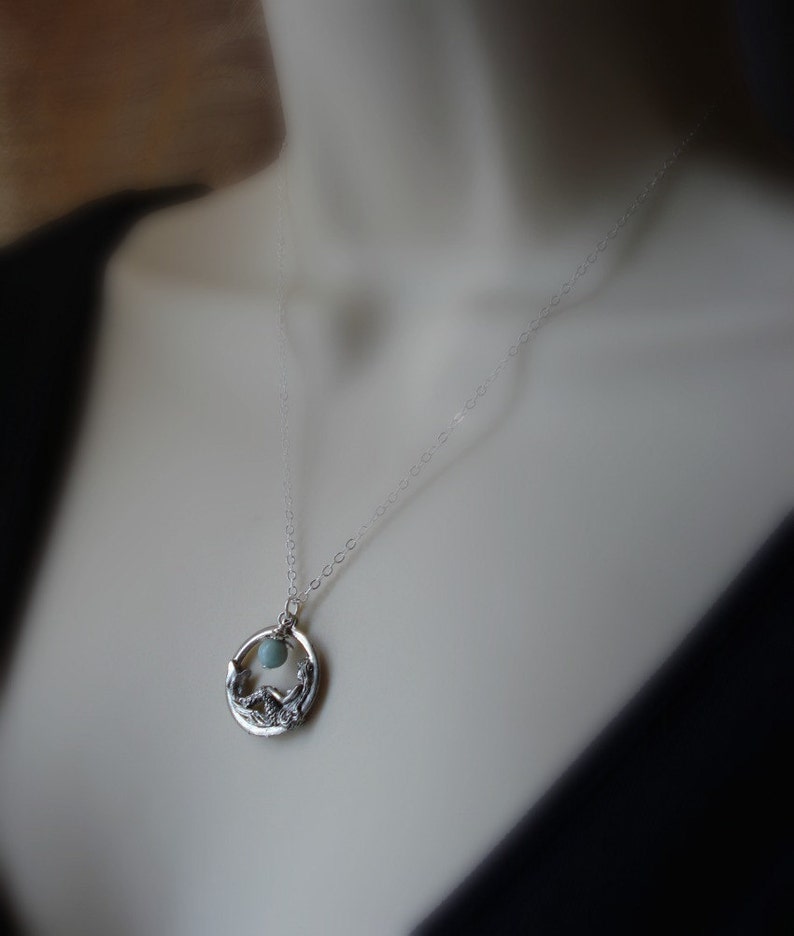 May include: A silver necklace with a delicate chain and a pendant featuring a circular design with a small blue gemstone in the center. The pendant is detailed with a stylized floral motif.