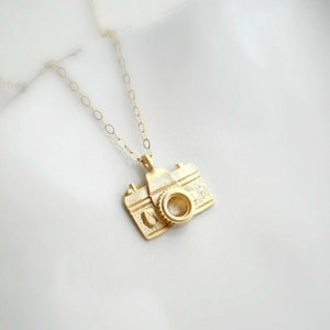 May include: A gold camera charm necklace. The camera charm is detailed and has a lens.