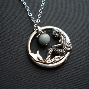 May include: Silver necklace with a mermaid pendant and a light blue gemstone. The mermaid is sitting on a wave and holding a pearl.
