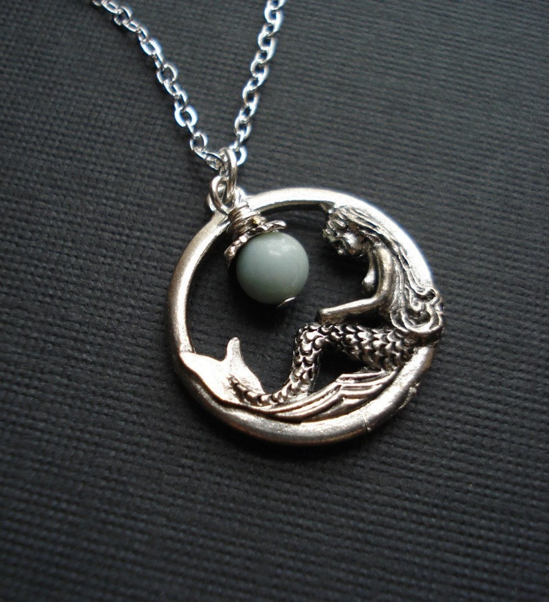 May include: Silver necklace with a mermaid pendant and a light blue gemstone. The mermaid is sitting on a wave and holding a pearl.