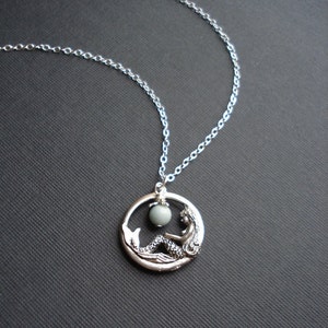 May include: A silver necklace with a circular pendant featuring a mermaid sitting inside a circle with a light green gemstone.