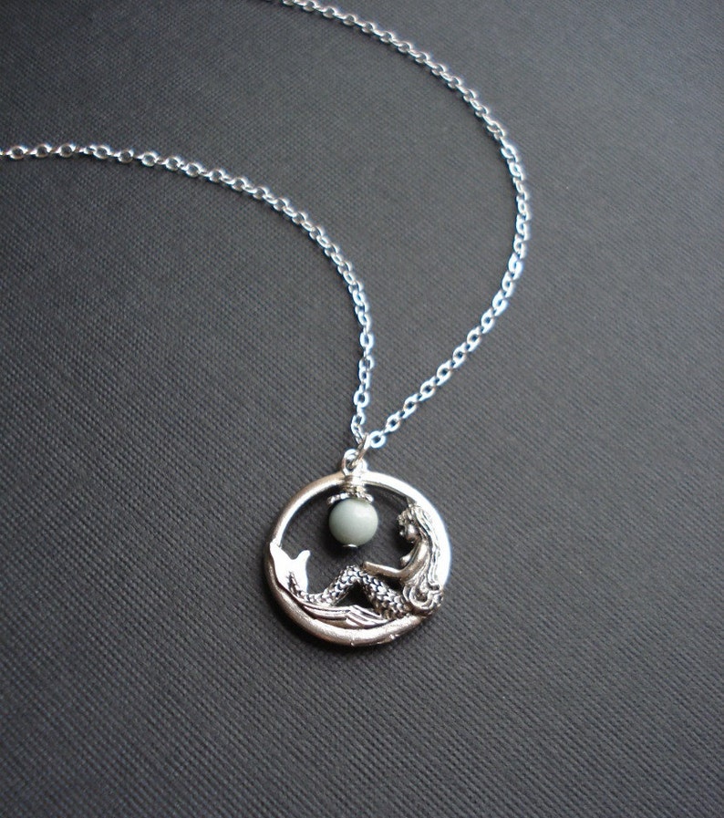 May include: A silver necklace with a circular pendant featuring a mermaid sitting inside a circle with a light green gemstone.
