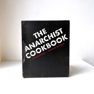 Könnte beinhalten: Ein schwarzes Buch mit dem Titel "The Anarchist Cookbook" in weißer, fetter, serifenloser Schrift. Der Name des Autors, William Powell, und der Autor der Einführung, Peter Bergman, sind ebenfalls auf dem Cover aufgeführt.
