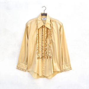Op de afbeelding: Een vintage beige shirt met lange mouwen en ruches aan de voorkant en manchetten. Het shirt heeft een puntkraag en een knoopsluiting. De ruches zijn in een donkerdere tint beige, wat een decoratief element aan het kledingstuk toevoegt.