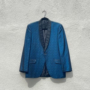 May include: A blue blazer with a black lapel and a single button closure. The jacket is hanging on a black hanger against a textured white wall. The blazer has two front pockets.