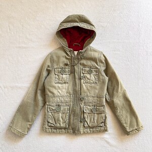 May include: An olive green hooded jacket with a red interior lining. The jacket features four front pockets with button closures, a drawstring hood, and buttoned cuffs. The jacket is made of a durable fabric.