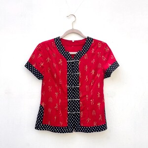 May include: Red short-sleeved top with gold script and black and white trim. The top has a V-neck and a series of white buttons down the front. The trim is a black and white pattern. The top is hanging on a wooden hanger.