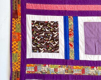 May include: A purple patchwork quilt featuring a range of fabrics and designs, including a brown fabric with white birds, a blue fabric with paisley patterns, and a purple fabric with a floral pattern. The quilt is edged with a yellow and orange fabric with geometric patterns.