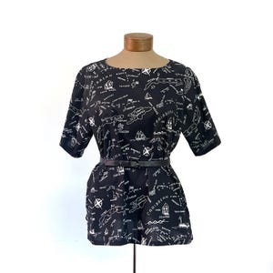 May include: Black short-sleeved dress with a Caribbean map print in white. The dress has a round neckline and is cinched at the waist with a black belt. The map design includes illustrations of sailboats and compasses.