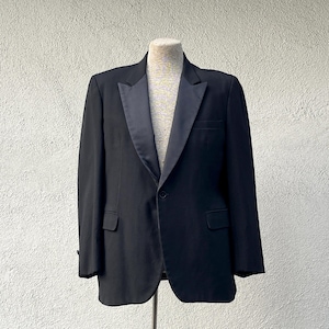 May include: A black tuxedo jacket with a satin lapel, displayed on a mannequin. The single-breasted jacket has a one-button closure and two front pockets. The formal wear is suitable for special occasions.