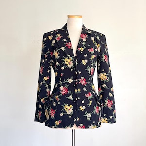 May include: A black blazer with a floral pattern in red, yellow, and white. The blazer has a notched collar, two rows of buttons, and two front pockets. The garment is displayed on a mannequin.