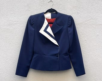 May include: A navy blue blazer with a white geometric design on the lapel and collar. The blazer has long sleeves, a single-button closure, and a red interior lining. The jacket is hanging on a black hanger against a light gray wall.