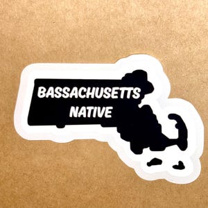 May include: A sticker with a white border, displaying a black outline of Massachusetts. The words "MASSACHUSETTS NATIVE" are in white within a black rectangle. The sticker is on a brown background.