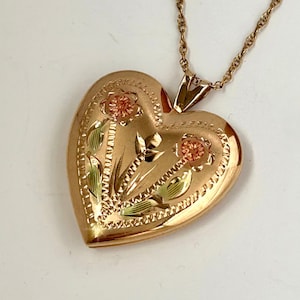 May include: A heart-shaped locket pendant crafted from rose gold, featuring engraved floral details with green and pink gold accents. The locket is suspended from a delicate matching rose gold rope chain, showcasing a polished finish.