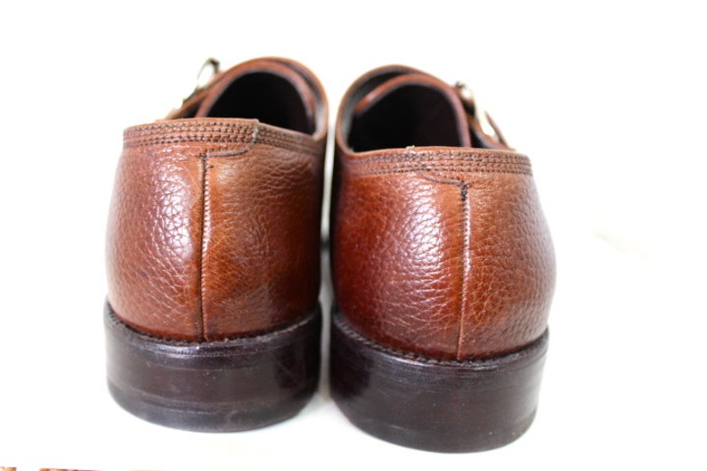 May include: A pair of brown leather monk strap shoes with a textured finish. The shoes have a dark brown sole and a buckle closure.