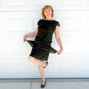 May include: A woman in a black sequined dress with short sleeves poses in front of a white garage door.