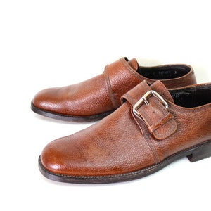 May include: A pair of brown leather monk strap shoes with a buckle closure. The shoes have a textured leather upper and a dark brown sole.