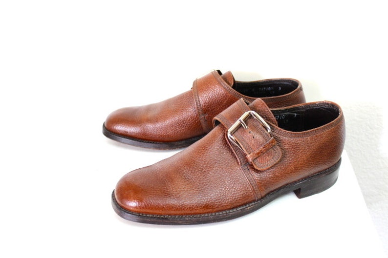 May include: A pair of brown leather monk strap shoes with a buckle closure. The shoes have a textured leather upper and a dark brown sole.