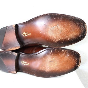 May include: Close-up of the brown leather soles of a pair of shoes. The soles are worn and have a slight sheen. The shoes are made by "Barker England".