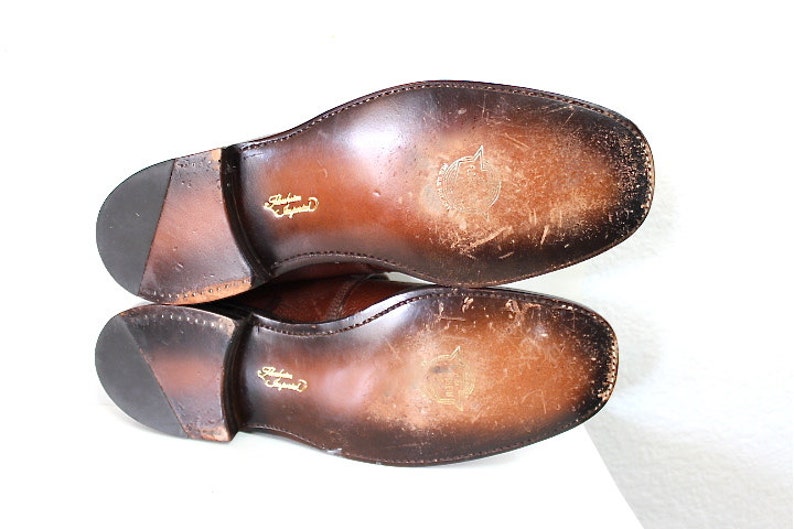 May include: Close-up of the brown leather soles of a pair of shoes. The soles are worn and have a slight sheen. The shoes are made by "Barker England".