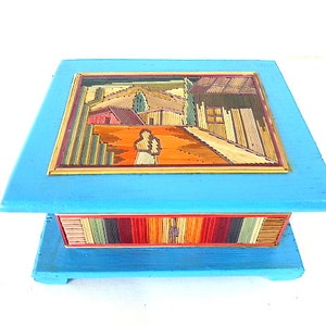 May include: A blue wooden box with a colorful, stick-like mosaic design on the top and bottom. The mosaic depicts a landscape scene with houses and trees.