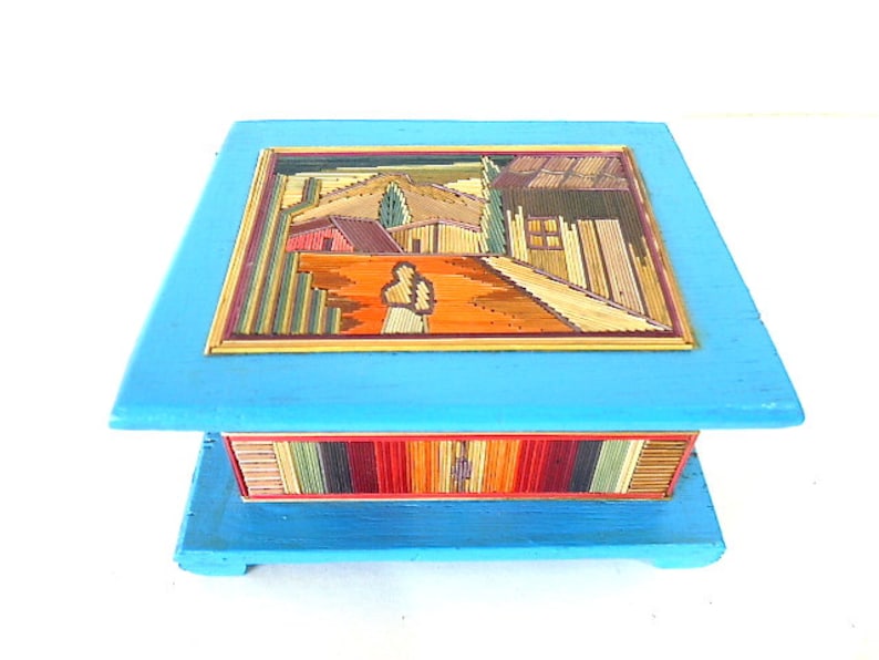May include: A blue wooden box with a colorful, stick-like mosaic design on the top and bottom. The mosaic depicts a landscape scene with houses and trees.