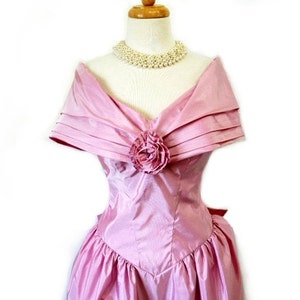 May include: A pink satin dress with a large flower detail at the neckline. The dress has a sweetheart neckline and off-the-shoulder straps. The dress is fitted at the waist and flows out into a full skirt.