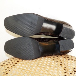 Op de afbeelding: Een paar bruine, getextureerde leren schoenen met vierkante neuzen en blokhakken. De zolen zijn zwart met een textuurpatroon. De schoenen liggen op een lichtgekleurd, geweven oppervlak.