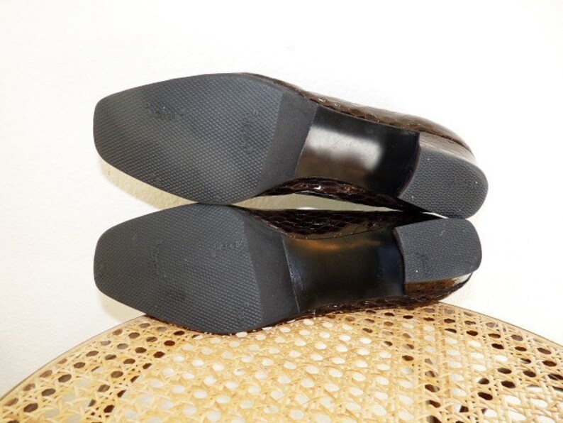 Op de afbeelding: Een paar bruine, getextureerde leren schoenen met vierkante neuzen en blokhakken. De zolen zijn zwart met een textuurpatroon. De schoenen liggen op een lichtgekleurd, geweven oppervlak.