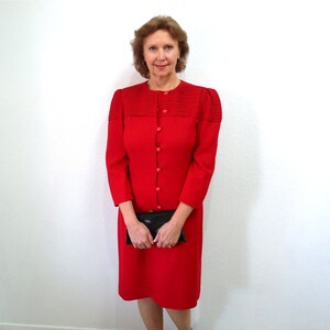 May include: A bright red dress with long sleeves and a button-up front. The dress features a round neckline and a knee-length hem. The shoulders have a pleated design. A black clutch is held in front of the dress.