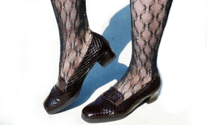 Op de afbeelding: Een paar donkerbruine, leren loafers met een textuur en een lage hak. De schoenen worden gecombineerd met zwarte, patroon, glinsterende kousen. De loafers hebben een klassiek ontwerp met een vierkante neus en een decoratieve band.