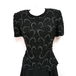 May include: Black beaded top with short sleeves. The top features intricate floral patterns and a fitted waist. The garment is displayed on a white mannequin, showcasing its design and texture. The top is part of a formal wear collection.