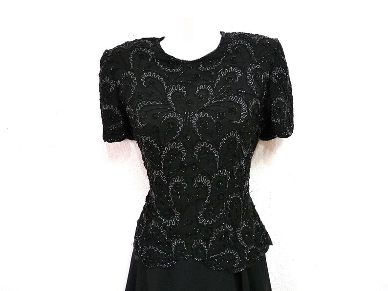 May include: Black beaded top with short sleeves. The top features intricate floral patterns and a fitted waist. The garment is displayed on a white mannequin, showcasing its design and texture. The top is part of a formal wear collection.