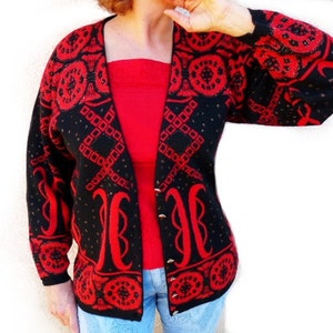 Può includere: Un cardigan nero con un motivo geometrico rosso e bottoni dorati. Il maglione aperto presenta un design audace con forme a diamante e circolari. Le maniche e i bordi sono rifiniti con lo stesso motivo rosso. Il cardigan è indossato sopra un top rosso e jeans blu.