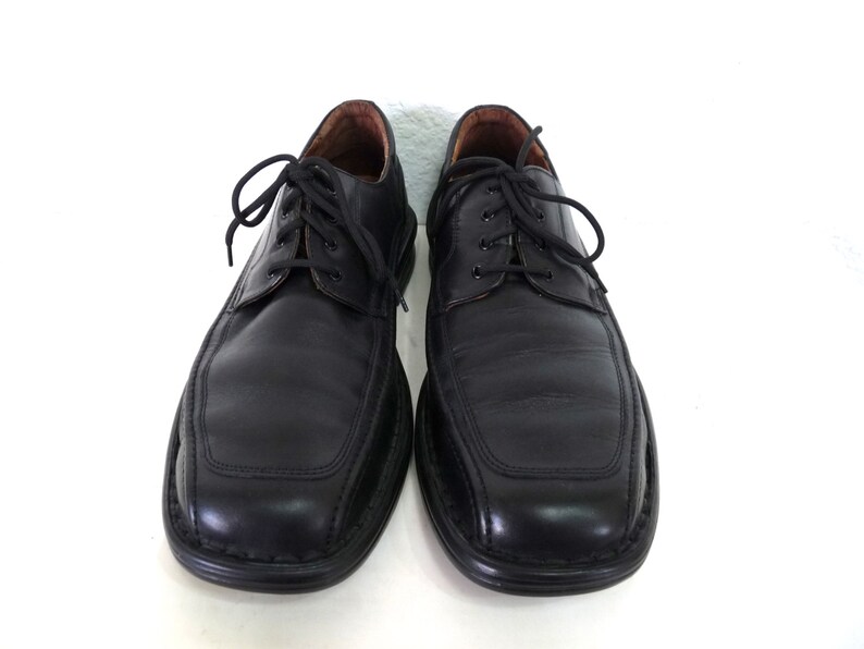 May include: A pair of black leather lace-up shoes with a rounded toe and a stitched design on the front. The shoes have a black rubber sole.
