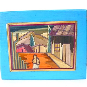 May include: A framed artwork depicting a rural scene with houses and a figure in the foreground. The artwork is made of straw and is set against a blue background.