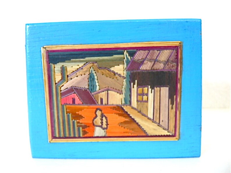 May include: A framed artwork depicting a rural scene with houses and a figure in the foreground. The artwork is made of straw and is set against a blue background.